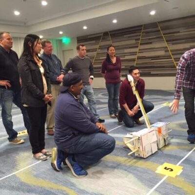 A group of participants indoors discussing their catapult design as part of the Catapult to Success team building activity, focusing on teamwork, strategy, and collaborative problem-solving in a competitive setting. thumbnail