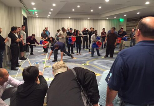 Participants in an indoor setting engaging in a competitive team building activity with catapults, buckets, and various objects, demonstrating teamwork, strategy, and coordination during the Catapult to Success event.