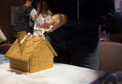 Close-up of a person decorating a gingerbread house by piping white icing on the roof and adding peppermint candies, during a holiday-themed team building event.