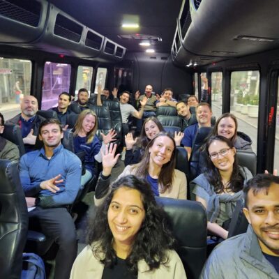 A group of smiling participants sits inside a bus, waving at the camera during a Do Good Bus team building event. The participants are dressed casually, ready to engage in a community service activity. thumbnail