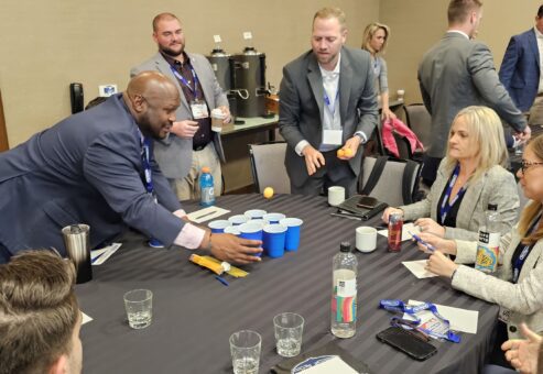 Group of professionals participating in a corporate team building event. One man arranges blue cups on the table while others watch and engage in discussion.