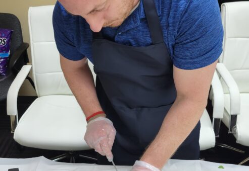 A man in a blue shirt and black apron is carefully chopping an onion at a table during a team building chili cook-off event. He is wearing disposable gloves, and there are herbs, chopped vegetables, and a small bowl with ingredients on the table in front of him.