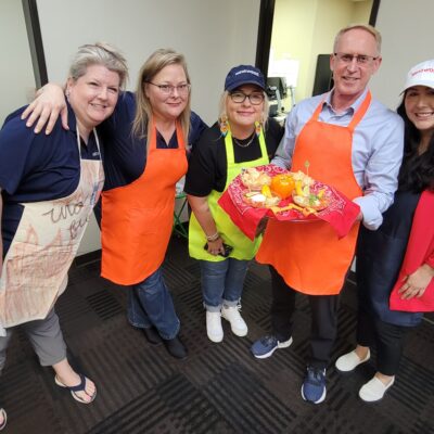 A group of five people smiling and posing, holding a tray of food. They are participating in a chili cook-off team building activity. Three of the participants wear bright aprons, one in orange and one in lime green. The man in the center is holding a red tray with an arrangement of colorful vegetables and food items. thumbnail