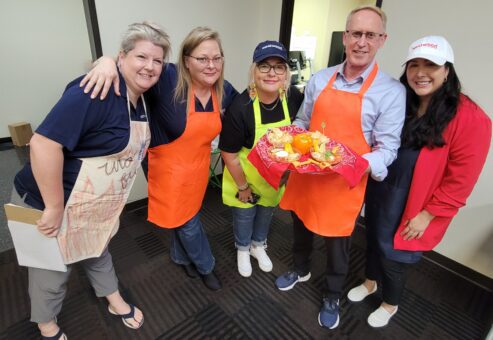 A group of five people smiling and posing, holding a tray of food. They are participating in a chili cook-off team building activity. Three of the participants wear bright aprons, one in orange and one in lime green. The man in the center is holding a red tray with an arrangement of colorful vegetables and food items.