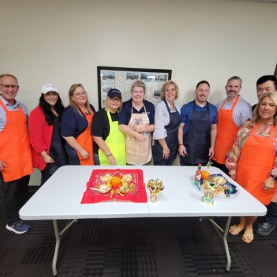A group of individuals stands together, smiling, behind a table at a team building chili cook-off event. Some are wearing brightly colored aprons in shades of orange, blue, and green. The table in front of them displays two creatively presented chili dishes, along with small golden trophies. thumbnail