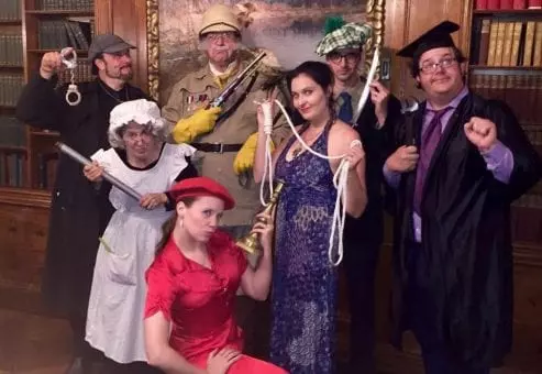 A group of seven participants pose in costume for a themed Murder Mystery Dinner team building event. Each person is dressed as a unique character, holding props that represent classic murder mystery items, such as a candlestick, rope, and revolver.