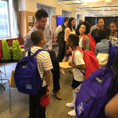 A team building participant interacts with children during a Tools for Schools CSR event, as they receive brightly colored backpacks filled with school supplies. thumbnail