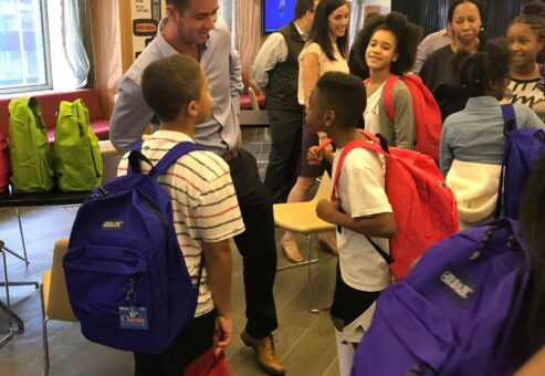 A team building participant interacts with children during a Tools for Schools CSR event, as they receive brightly colored backpacks filled with school supplies.