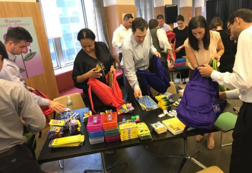 Corporate team members packing backpacks with school supplies during a csr team building event. The table is filled with notebooks, pencils, and other educational materials as participants work together to prepare donations, emphasizing teamwork and giving back to the community.