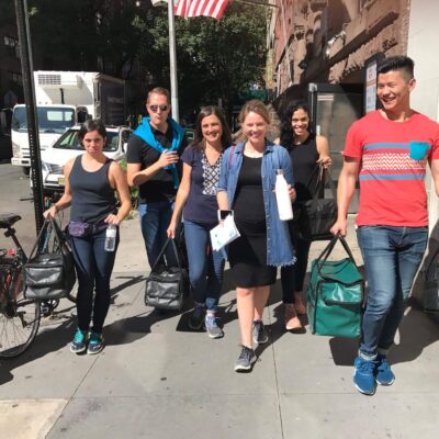 A group of six people walking down a city sidewalk, smiling and carrying insulated bags. They are engaged in a team building activity, possibly delivering meals or supplies. thumbnail