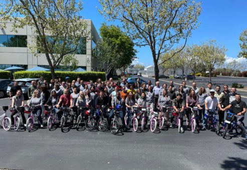 Large group of participants posing outdoors with assembled bikes during a Charity Bike Build event. The group stands proudly behind the rows of bikes, enjoying the sunny day and the sense of accomplishment after working together for a charitable cause.