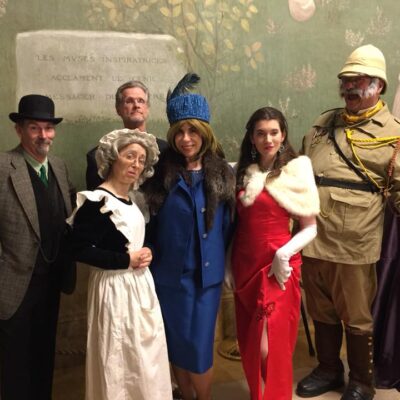 A group of actors dressed in period costumes for a Murder Mystery Dinner event. The characters include a man in a detective's coat and hat, a maid in a traditional white apron, a woman in a blue dress and fur hat, a glamorous woman in a red gown and fur shawl, and an explorer wearing a safari outfit and yellow gloves. thumbnail