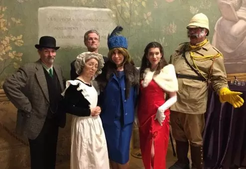 A group of actors dressed in period costumes for a Murder Mystery Dinner event. The characters include a man in a detective's coat and hat, a maid in a traditional white apron, a woman in a blue dress and fur hat, a glamorous woman in a red gown and fur shawl, and an explorer wearing a safari outfit and yellow gloves.