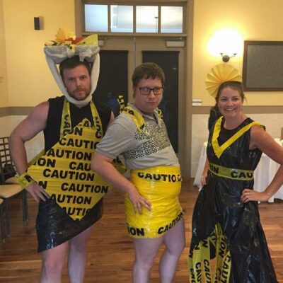 Three individuals pose confidently at a team building event, showcasing their creative outfits made from 'Caution' tape and other recyclable materials. The person on the left is wearing a black and yellow outfit, with a decorative headpiece made from caution tape. The person in the middle is also adorned in a yellow caution-tape skirt and a gray top. The individual on the right is dressed in a black and yellow dress with a matching headpiece, all made from caution tape and other materials. The mood is playful, and the group looks proud of their fashion designs. thumbnail