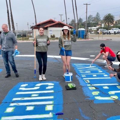 A group of volunteers is painting a large parking lot mural in bold blue and white letters. Two women in the center hold long paint rollers while smiling, and two other volunteers are crouched down applying paint. Palm trees and a beach setting are visible in the background, along with some vehicles parked nearby. Everyone appears to be enjoying the outdoor charitable Do Good Bus team building program. thumbnail