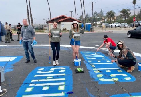 A group of volunteers is painting a large parking lot mural in bold blue and white letters. Two women in the center hold long paint rollers while smiling, and two other volunteers are crouched down applying paint. Palm trees and a beach setting are visible in the background, along with some vehicles parked nearby. Everyone appears to be enjoying the outdoor charitable Do Good Bus team building program.