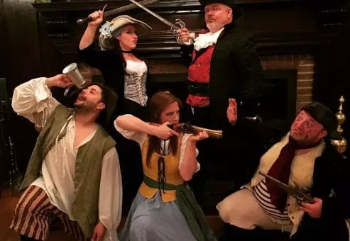 A group of actors in pirate-themed costumes pose dramatically for a Murder Mystery Dinner event. The characters include a pirate captain in a black hat and red vest, a woman dressed as a swashbuckler holding a sword, another woman in a long blue and yellow dress aiming a musket, and two additional pirates—one with a mug and the other with a flintlock pistol. They stand in front of an elegant fireplace, creating a lively and adventurous scene.