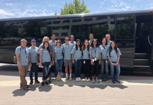 A group of fourteen people standing in front of a large black bus, all wearing matching light blue t-shirts, participating in a team building event. The individuals are smiling and standing close together, indicating camaraderie and a shared purpose.