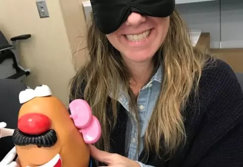 A woman smiling and holding a Mr. Potato Head toy while wearing a black blindfold. She is participating in a fun activity as part of the Toys for Tykes team building event, where participants assemble a toy blindfolded as a challenge.