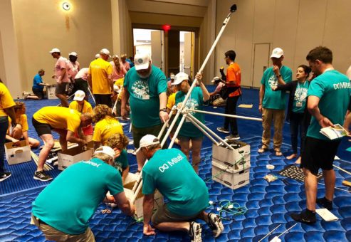 Multiple teams in brightly colored shirts collaborating on building catapults indoors during the Catapult to Success team building activity, showcasing teamwork, creativity, and hands-on construction skills in a lively group setting.