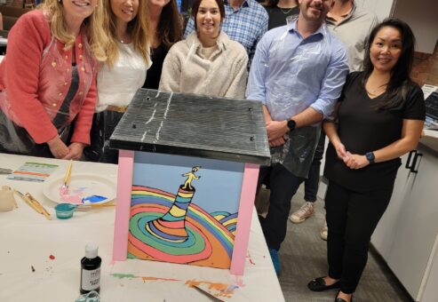 Team proudly poses with their completed Little Free Library during a team building event. The vibrant library features a whimsical design, showcasing teamwork, creativity, and a shared effort to inspire community literacy through the creation of a colorful and engaging book-sharing structure.