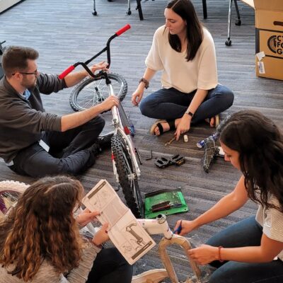 A group of four people sitting on the floor assembling a bicycle during a Charity Bike Build event. They are carefully reading instructions and working collaboratively, with bike parts and tools spread around them. thumbnail