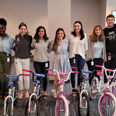 A group of smiling participants standing behind a row of assembled children's bicycles during a Charity Bike Build event. The bikes are pink and blue with training wheels, and the group appears happy with their completed team project. thumbnail