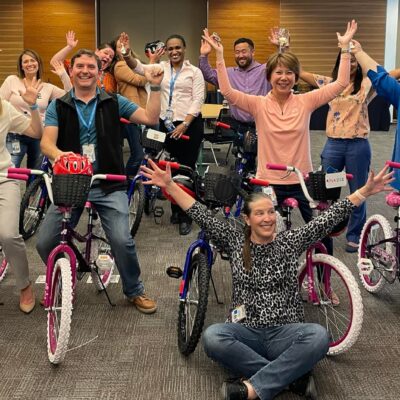 A group of excited participants proudly displaying their completed bicycles during a Charity Bike Build event. They stand behind and beside the assembled bikes with their arms raised in celebration. thumbnail