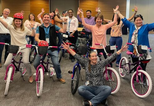 A group of excited participants proudly displaying their completed bicycles during a Charity Bike Build event. They stand behind and beside the assembled bikes with their arms raised in celebration.