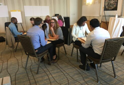 Small groups of participants engage in one-on-one discussions while holding wooden boards during a board-breaking team building activity focused on overcoming personal barriers.