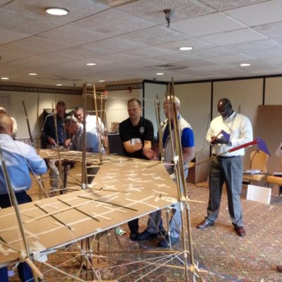 Participants in the Bridging the Divide team building activity gather around a large, collaboratively built cardboard bridge, assessing its structure and stability. thumbnail