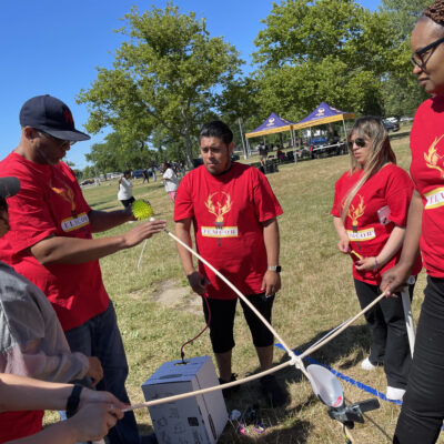 A team of participants in red shirts working together outdoors on constructing a catapult during the Catapult to Success team building activity, showcasing collaboration, creativity, and strategic planning. thumbnail