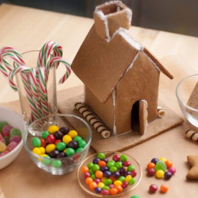 A partially constructed gingerbread house sits on a wooden table surrounded by bowls of colorful candies, including gumdrops, Skittles, and candy canes. The gingerbread house features a tall roof and chimney, with wafer sticks used as decorative accents along the base. The scene is ready for further decoration during the team building event. thumbnail