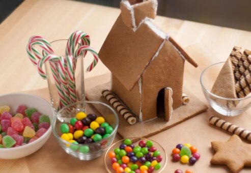 A partially constructed gingerbread house sits on a wooden table surrounded by bowls of colorful candies, including gumdrops, Skittles, and candy canes. The gingerbread house features a tall roof and chimney, with wafer sticks used as decorative accents along the base. The scene is ready for further decoration during the team building event.