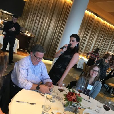 A scene from a Murder Mystery Dinner team building event, where a female actor, holding a microphone, engages with participants seated at a round table. One man listens attentively while another woman smiles. The event appears lively and interactive as the actor involves the audience in the storyline. thumbnail