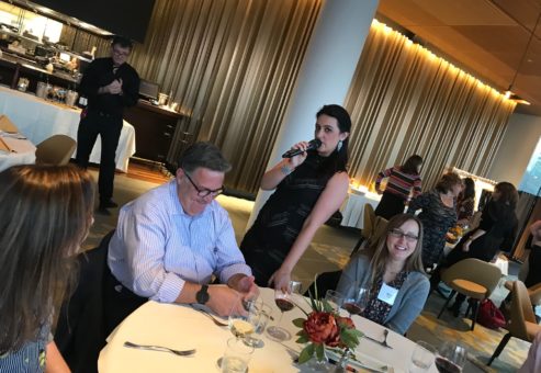 A scene from a Murder Mystery Dinner team building event, where a female actor, holding a microphone, engages with participants seated at a round table. One man listens attentively while another woman smiles. The event appears lively and interactive as the actor involves the audience in the storyline.