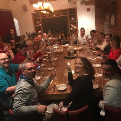 A large group of people gathered around a long wooden table in a warmly lit dining room for a Murder Mystery Dinner event. Each person is holding a glass of wine, smiling, and raising their glass in a celebratory toast. The table is set with elegant dinnerware and a centerpiece featuring a decorative cake. The atmosphere is festive, with participants ready to enjoy both the meal and the interactive murder mystery experience. thumbnail