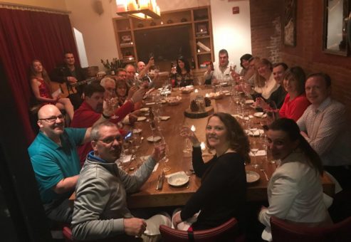 A large group of people gathered around a long wooden table in a warmly lit dining room for a Murder Mystery Dinner event. Each person is holding a glass of wine, smiling, and raising their glass in a celebratory toast. The table is set with elegant dinnerware and a centerpiece featuring a decorative cake. The atmosphere is festive, with participants ready to enjoy both the meal and the interactive murder mystery experience.