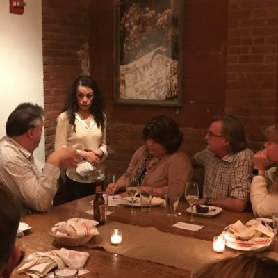 A group of four people sitting around a dimly lit dinner table engaged in conversation, with a female actor standing and looking at one of the seated individuals. The setting suggests an intimate dining experience, part of a Murder Mystery Dinner event, as the group interacts with the actor in the unfolding mystery. thumbnail