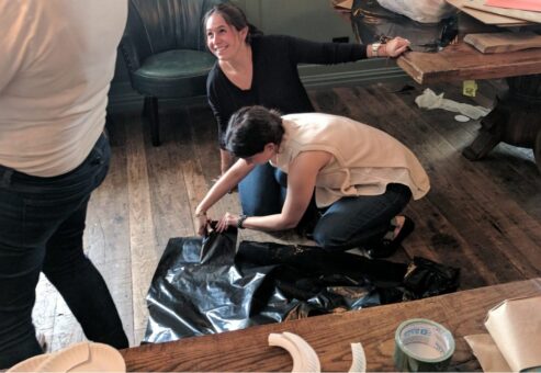 Two women engaged in a crafting activity for a team building event. One is kneeling on the wooden floor, cutting or adjusting a piece of black plastic material, while the other woman leans on a nearby table, smiling and watching.