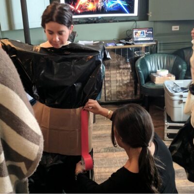 Participants collaborate during a Corporate Project Runway team building fashion activity. A woman stands in the center while others assist in constructing a garment out of black plastic material and cardboard. One person is adjusting the fit by attaching materials around her waist. thumbnail