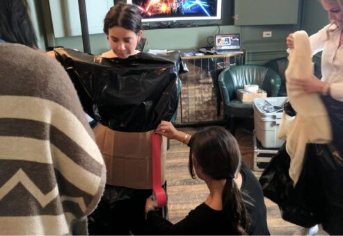 Participants collaborate during a Corporate Project Runway team building fashion activity. A woman stands in the center while others assist in constructing a garment out of black plastic material and cardboard. One person is adjusting the fit by attaching materials around her waist.