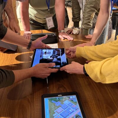 Participants in a 'Corporate CSI' team building event gather around a table, using iPads to examine digital evidence and clues as they work collaboratively to solve the mystery. thumbnail