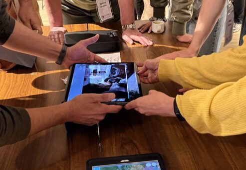 Participants in a 'Corporate CSI' team building event gather around a table, using iPads to examine digital evidence and clues as they work collaboratively to solve the mystery.