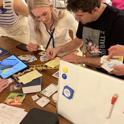 Team members actively engaged in solving a mystery during the 'Corporate CSI' team building event. Participants are examining evidence cards, taking notes, and using digital tools on tablets to analyze clues. thumbnail