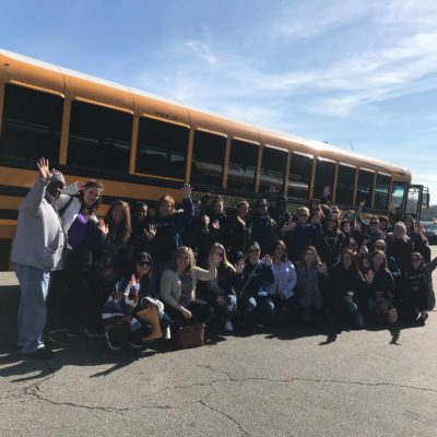 A large group of people poses in front of a yellow school bus on a sunny day, many of them waving at the camera. They are outdoors in a parking lot, with clear skies above, creating a sense of enthusiasm and camaraderie. The group appears to be preparing for or returning from a volunteer outing, part of a team building event. The mix of smiles and waves highlights the positive energy and collaborative spirit of the group. thumbnail