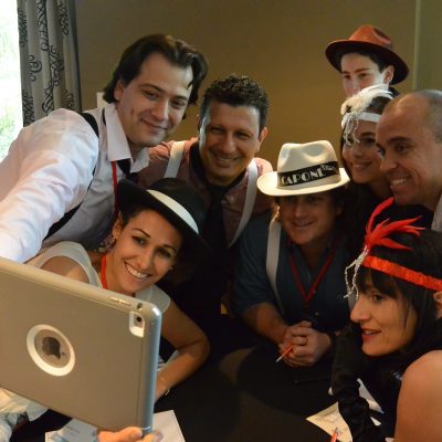 A team dressed in 1920s gangster-themed outfits gathers for a selfie during the high-energy Escape the Mob team building event, solving clues and creating their alibi. thumbnail