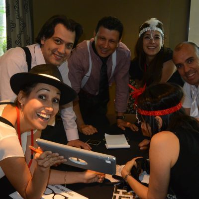 A group of participants in the Escape the Mob team building event works together, using a tablet to solve clues and track down the stolen diamonds while embracing their mobster-themed costumes. thumbnail