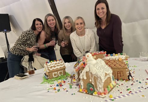 Five smiling participants gather around a table displaying their decorated gingerbread houses during a team building event. The gingerbread houses are intricately adorned with icing, candy, and festive decorations, including a snowman figure on one of the houses.
