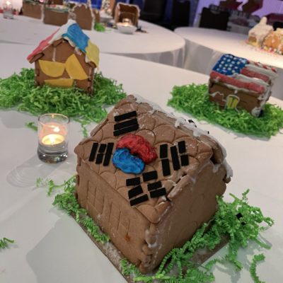 A gingerbread house decorated with icing and black licorice pieces, featuring the South Korean flag symbol in the center of the roof. The house is placed on a white table with green decorative grass around it, alongside a small lit candle. Other uniquely decorated gingerbread houses are visible in the background as part of a team building event. thumbnail